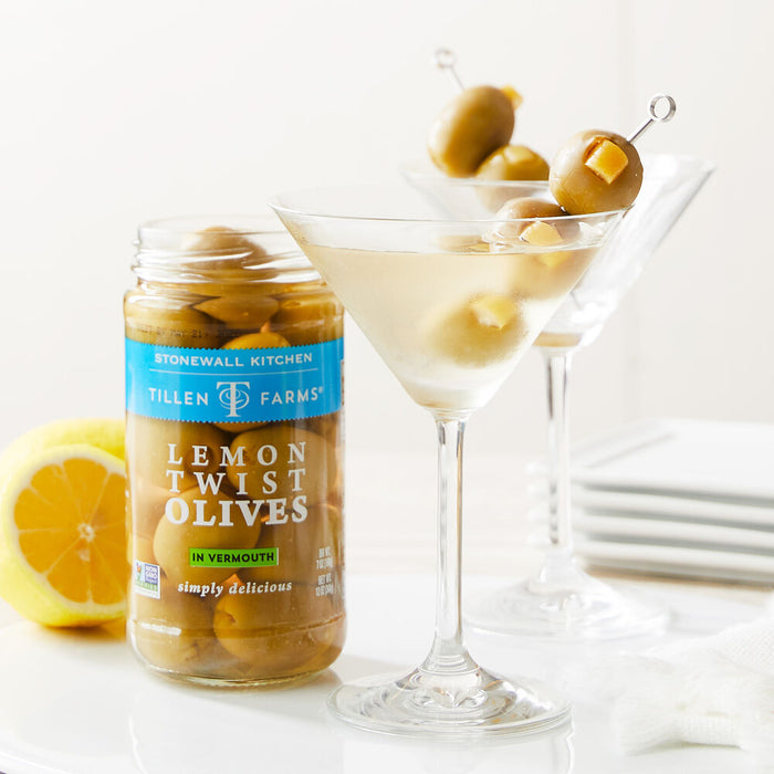 Jar of Tillen Farms Lemon Twist Olives next to a Martini cocktail glass with olives on a skewer on a white background