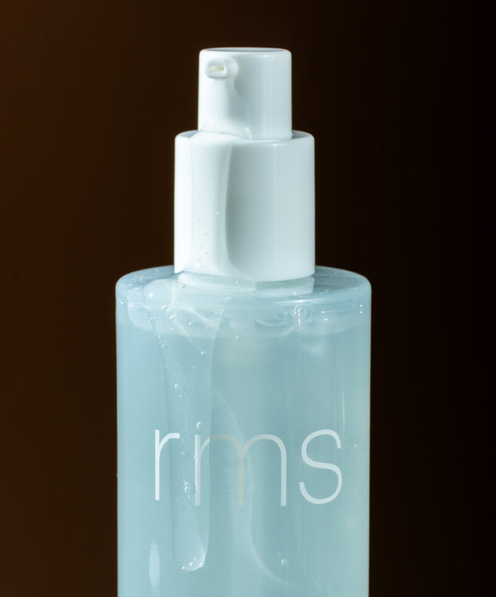 Clear blue cleansing gel in a transparent pump bottle labeled with RMS, showcasing the texture of Kakadu Clean Cleansing Gel against a dark background in clean beauty packaging.