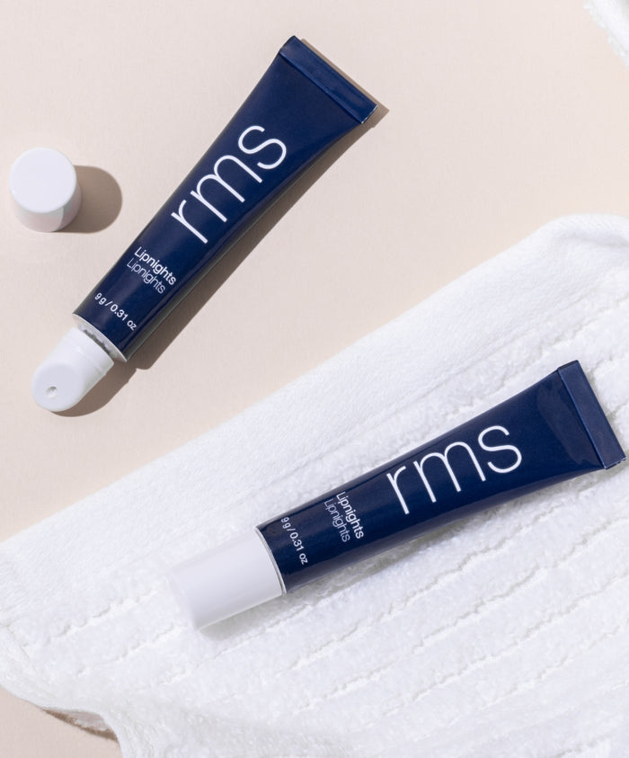 Two navy blue tubes of Lipnights Overnight Lip Mask with white caps, one open and one closed, displayed on a soft white textured cloth against a light background.