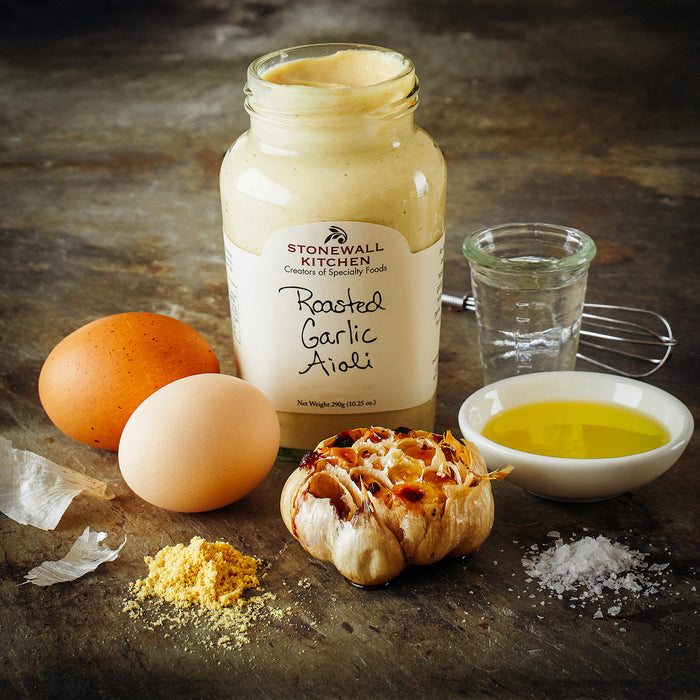 photo is an open jar of Stonewall Kitchen roasted Garlic aioli surrounded by the ingredients to make it eggs, garlic, oil, salt, and mustard powder