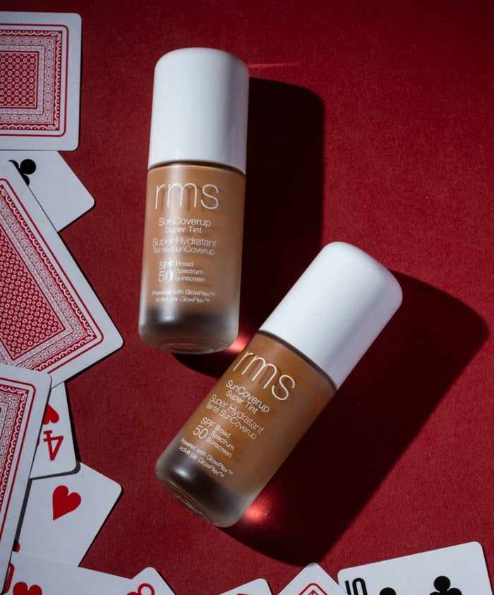 Two bottles of SunCoverup Super Tint Broad Spectrum SPF 50 Sunscreen by RMS Beauty placed on a red surface with scattered playing cards, highlighting clean beauty and sustainable packaging.